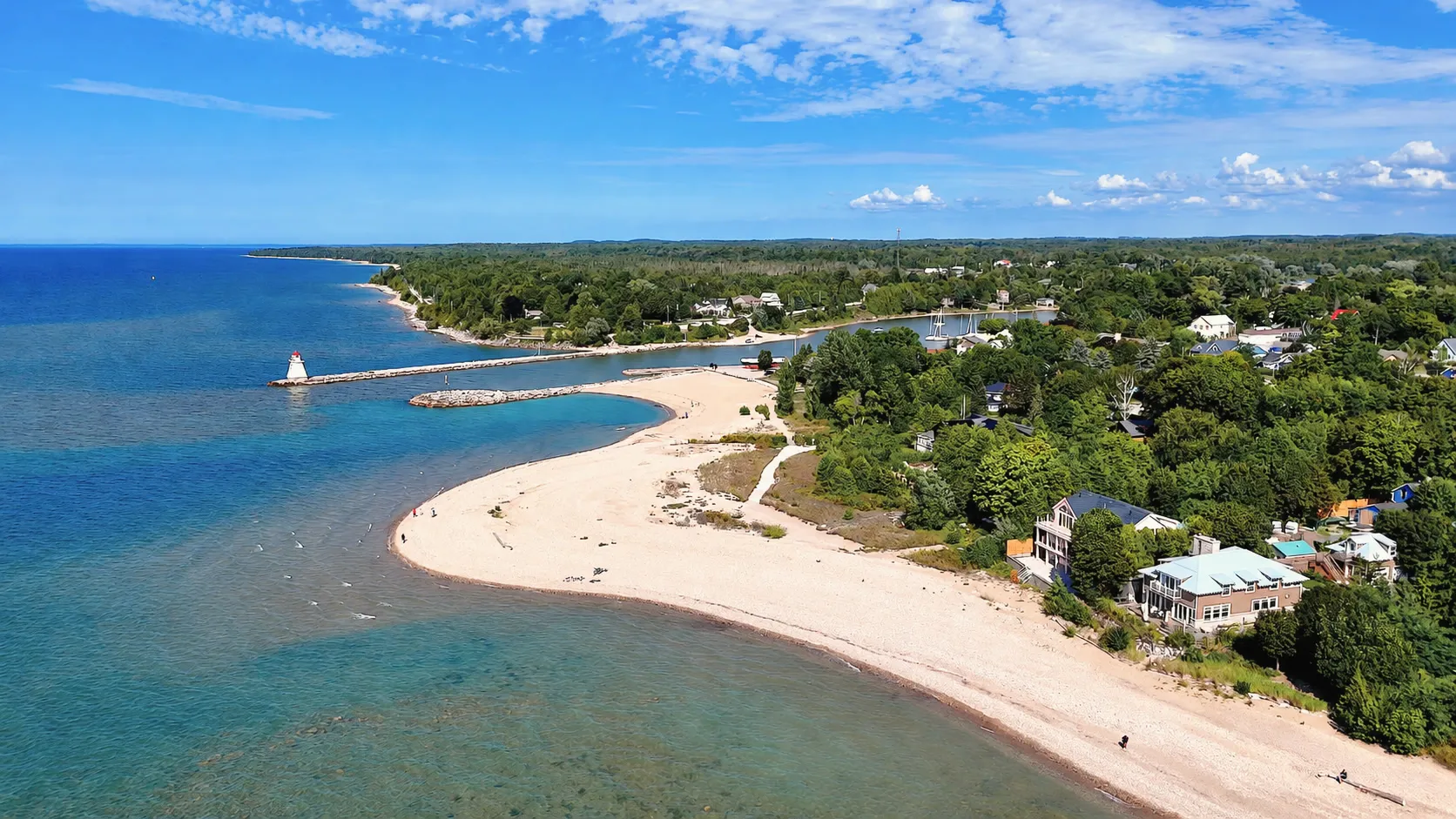 The beach from the air: a long curve of sand along Lake Huron, the breakwall and its range light at the river mouth, and homes in the trees behind