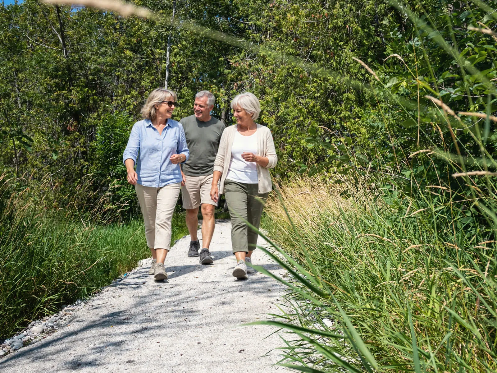 Three friends walking and talking on a gravel trail between tall grasses and trees