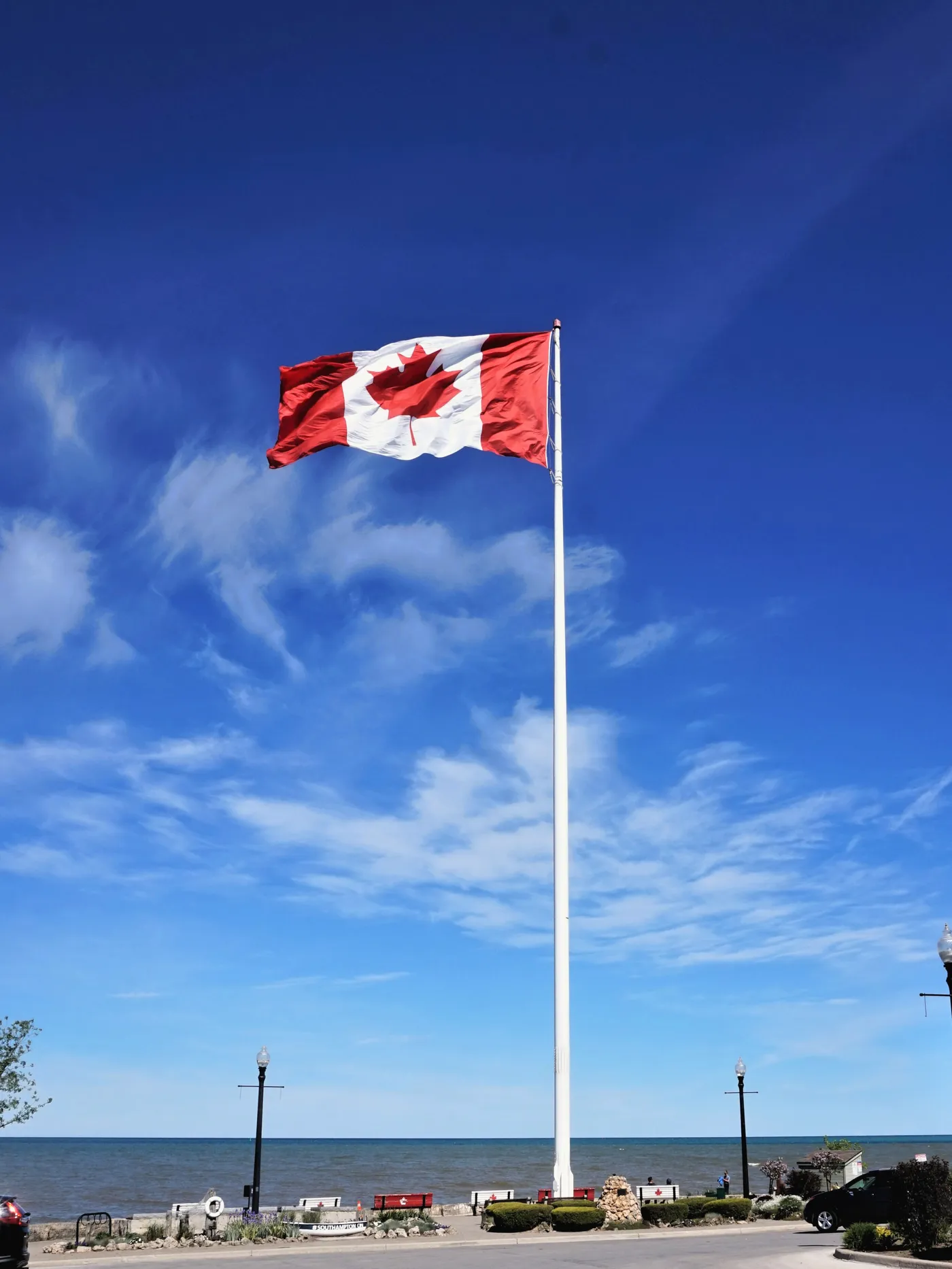 A large Canadian flag flying from a tall flagpole by the beach, with Lake Huron behind it