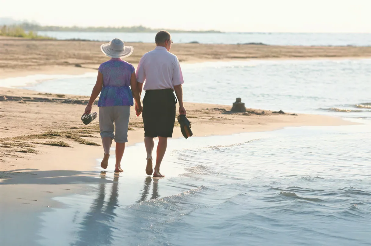 A couple walking barefoot along the water's edge, carrying their sandals